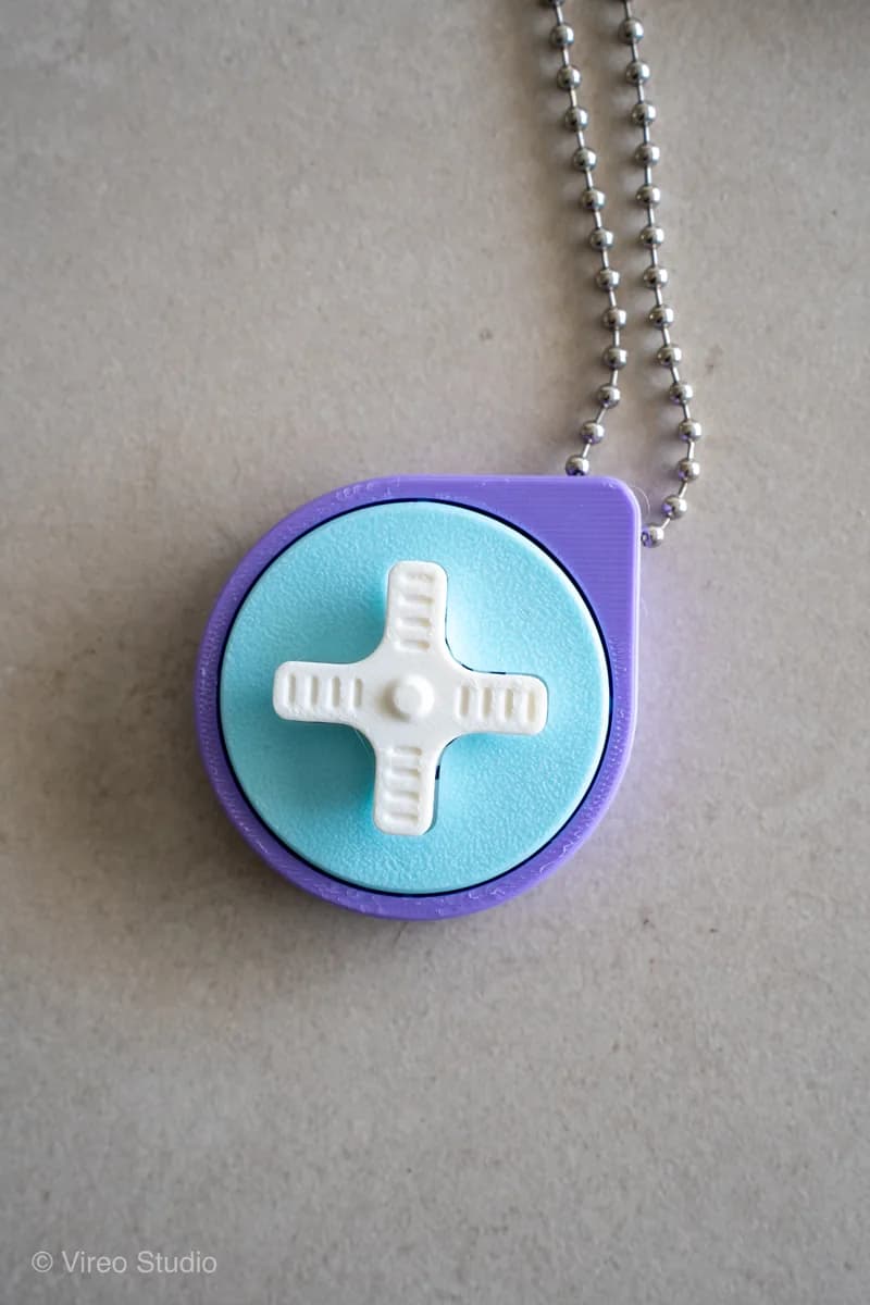 Retro game sleutelhanger van boven: lichtblauw rond vlak met paarse rand en een witte kruisvormige D-pad, aan een kogelkettinkje