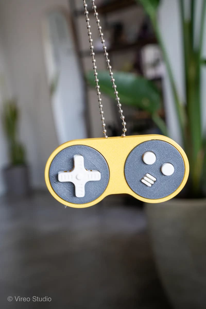 Game controller sleutelhanger: gele RetroSnap met donkergrijze vlakken, witte D-pad en lichte knopjes, aan een kogelkettinkje voor een plant