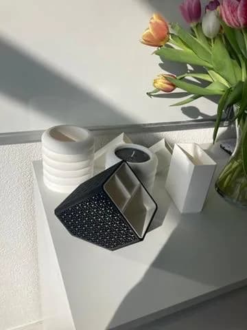 A black 3D-printed faceted vase beside white ceramics, with tulips in morning light.
