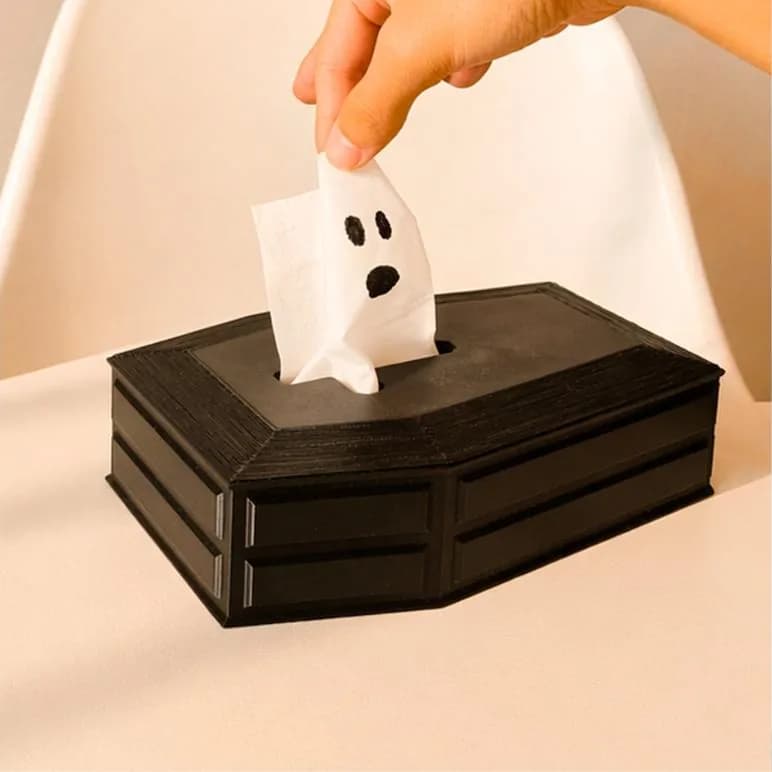 Doodskist-tissuebox – zwarte doodskist op tafel, een hand trekt er een tissue met spookgezichtje uit
