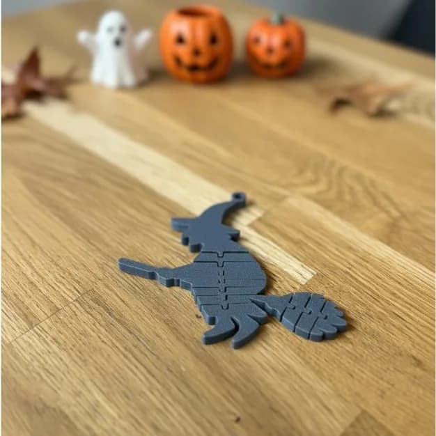 Heks op bezem – grijze flexi-heks op een houten tafel, met pompoenen en een spookje op de achtergrond